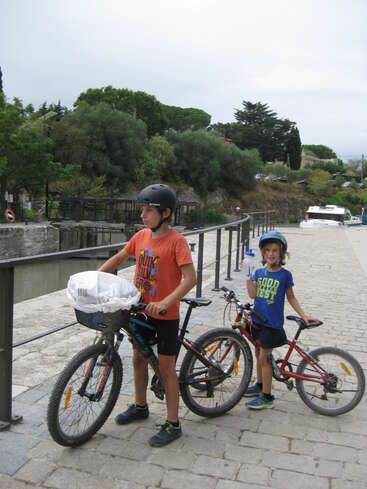 Duas crianças estão com suas bicicletas em uma passarela de pedra ao lado de um canal, usando capacetes e camisetas coloridas, com árvores e um céu nublado ao fundo.