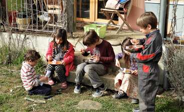 A imagem mostra um grupo de crianças sentadas em uma mesa, provavelmente comendo ou brincando, com alguns adultos presentes ao fundo, possivelmente supervisionando ou participando da atividade.