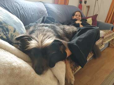 Uma mulher relaxa em um sofá, sorrindo com satisfação. Um cachorro fofo está deitado sobre as pernas dela, confortavelmente esticado e aconchegante, criando uma cena calorosa e pacífica.