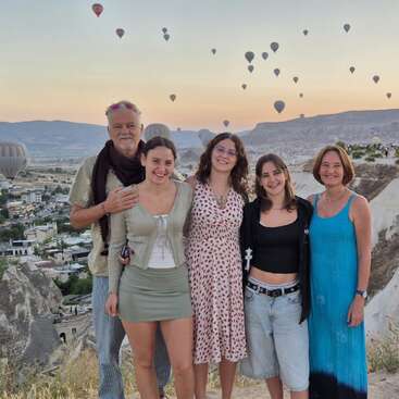 Un groupe souriant de cinq personnes pose en plein air au lever ou au coucher du soleil. Des montgolfières remplissent le ciel au-dessus d'un paysage pittoresque et rocheux et d'une ville charmante.
