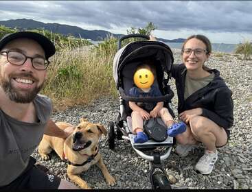 A happy family poses on a rocky beach: smiling man, woman, baby in stroller with emoji face, and cheerful dog. Mountains and water in background.