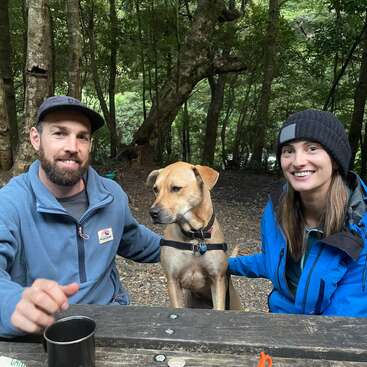 A man, woman, and brown dog sit together at a wooden picnic table in a forest, all wearing jackets and smiling, enjoying nature and each other's company.
