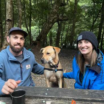 A man, woman, and brown dog sit together at a wooden picnic table in a forest, all wearing jackets and smiling, enjoying nature and each other's company.
