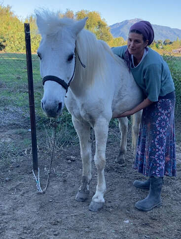 Uma mulher com uma saia estampada e botas está ao lado de um cavalo branco, abraçando-o gentilmente ao ar livre, perto de uma cerca com montanhas ao fundo.