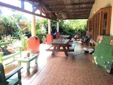 This cozy outdoor patio features wooden benches, picnic tables, hammocks, plants, a welcoming sign, and a shaded roof, creating a relaxing and inviting atmosphere.