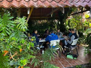 Six people sit around a white table under a tile-roofed gazebo surrounded by lush green plants, engaging in discussion or studying in a natural setting.