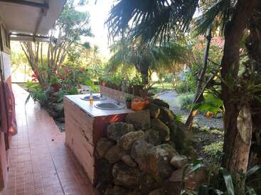 An outdoor sink surrounded by rocks and vibrant plants, located in a garden area next to a tiled walkway, under the shade of lush palm trees.