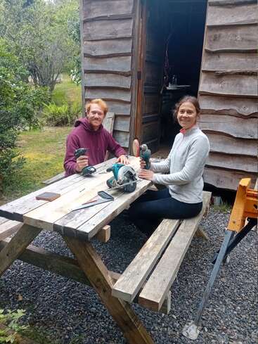 Zwei Menschen sitzen an einem hölzernen Picknicktisch und arbeiten lächelnd mit Elektrowerkzeugen. Sie stehen vor einem rustikalen Holzschuppen, umgeben von Grün und Bäumen.