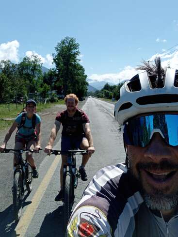 Drei Menschen genießen einen sonnigen Tag beim Radfahren auf einer offenen Straße. Sie sind von Bäumen und Bergen umgeben. Der vordere Radfahrer trägt eine Sonnenbrille und einen Helm und lächelt breit.