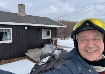Ein lächelnder Typ mit Helm macht ein Selfie vor einer kleinen schwarzen Hütte mit weißen Verzierungen, neben einem Schneemobil, umgeben von Schnee und winterlicher Landschaft.