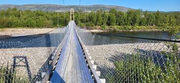 Eine schmale Hängebrücke mit Drahtgitterwänden führt über einen Fluss, umgeben von Bäumen und Bergen unter strahlend blauem Himmel, und lädt zu einem abenteuerlichen Ausflug ein.