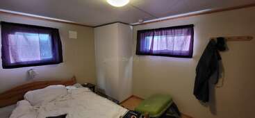 Das Bild zeigt ein kleines, einfaches Schlafzimmer mit einem ungemachten Bett, zwei Fenstern mit Vorhängen, einem weißen Kleiderschrank, einer Garderobe und minimaler Einrichtung. Gemütliche Atmosphäre.