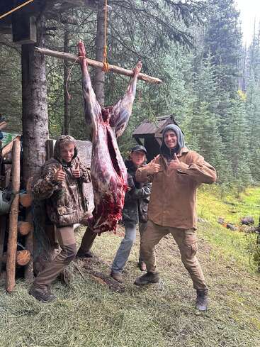 Drei Menschen in Outdoor-Kleidung posieren lächelnd und mit erhobenem Daumen neben einem frisch gehäuteten Tierkadaver, der an einer Holzkonstruktion in einem Wald hängt.