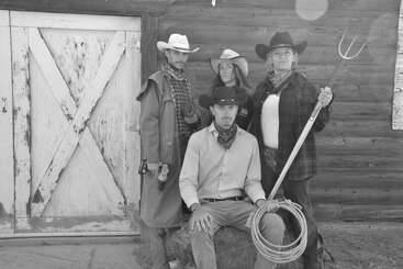 Vier als Cowboys und Cowgirls verkleidete Personen posieren ernsthaft vor einer rustikalen Scheune. Einer hält eine Mistgabel, ein anderer ein Seil, so dass eine Westernszene entsteht.