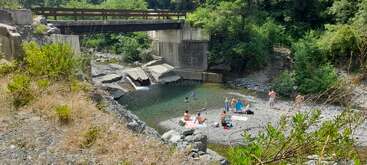 As pessoas relaxam e nadam em um rio rochoso sob uma pequena ponte, cercadas por árvores verdes exuberantes e pela natureza em um dia ensolarado, aproveitando o verão.