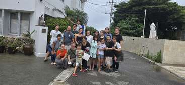 Un grupo de personas -adultos y niños- posan juntos frente a un edificio blanco en un día nublado, sonriendo y aparentando alegría sobre el pavimento mojado.