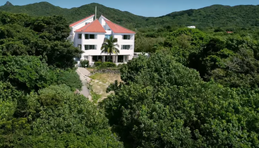 Una casa blanca con tejado rojo rodeada de frondosos árboles verdes y un denso bosque, con montañas visibles al fondo bajo un cielo azul y despejado.