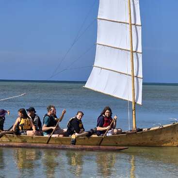 Six personnes portant des gilets de sauvetage font avancer un voilier en bois à voile blanche dans des eaux calmes et peu profondes, sous un ciel bleu clair, près du rivage.
