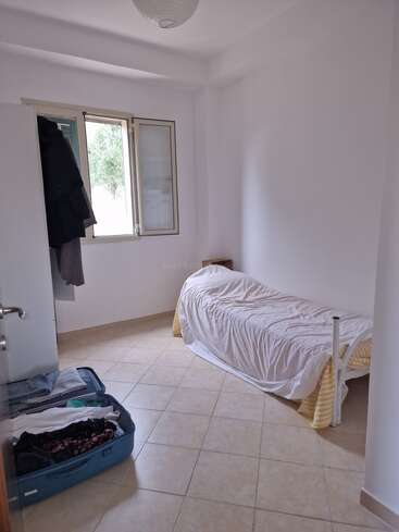 This is a small, simple bedroom with white walls, a single bed, an open window, wardrobe, coat hanging, tiled floor, and an open blue suitcase.