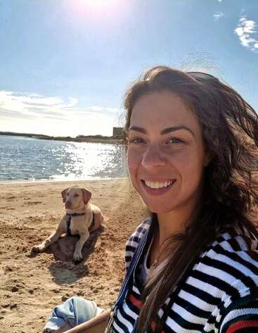 A woman smiles at the camera on a sunny beach, wearing a striped shirt. Behind her, a dog relaxes on the sand near the sparkling water.
