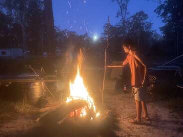 Ein junger Mensch steht in der Abenddämmerung an einem großen Lagerfeuer und hält einen Stock. Funken steigen in die Luft, umgeben von Bäumen und Campingausrüstung. Friedliche Atmosphäre.