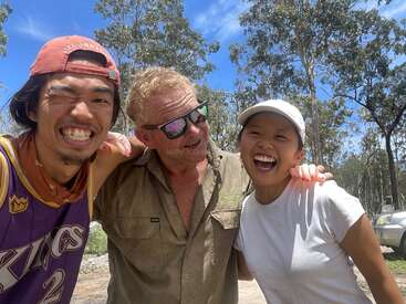 Drei Freunde posieren an einem sonnigen Tag im Freien und lächeln fröhlich. Hinter ihnen sind Bäume und der blaue Himmel zu sehen. Sie wirken glücklich, entspannt und genießen die Gesellschaft des anderen.