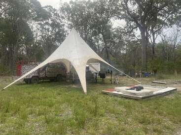 Ein dreieckiges weißes Zelt bedeckt einen Lagerplatz mit einem getarnten Fahrzeug, Stühlen und Tischen in einem grasbewachsenen Waldgebiet. In der Nähe liegt eine leere Matratze im Freien.