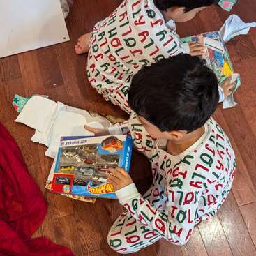 Zwei Kinder in festlichen Pyjamas öffnen aufgeregt Geschenke auf einem Holzboden. Eines hält ein Hot Wheels-Set, umgeben von zerrissenem Geschenkpapier und Weihnachtsstimmung.