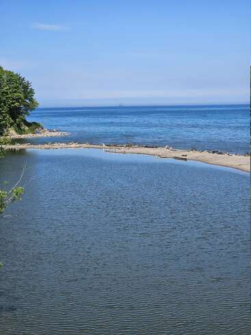 A serene scene shows a calm river meeting a blue lake beneath a clear sky, with sandy shores, distant trees, and distant ships on the horizon.