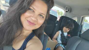A woman takes a cheerful selfie in a car, while a young child sits in a car seat in the back, looking down and relaxed.