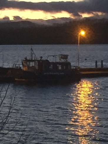A imagem mostra um barco em um corpo d'água ao pôr do sol, com um poste de luz e um céu nublado ao fundo. O barco é escuro e tem uma cabine.