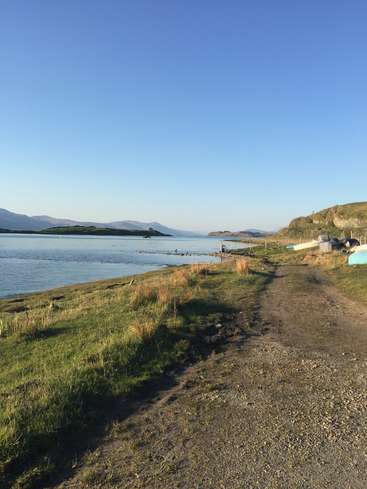 A imagem retrata uma cena costeira serena, com um caminho de terra, um terreno gramado e um corpo d'água, em meio a um céu azul claro e montanhas distantes.