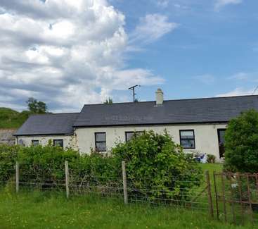 A imagem mostra uma casa de um andar com telhado cinza escuro e paredes brancas, cercada por uma cerca e vegetação exuberante, em um céu azul com nuvens brancas.