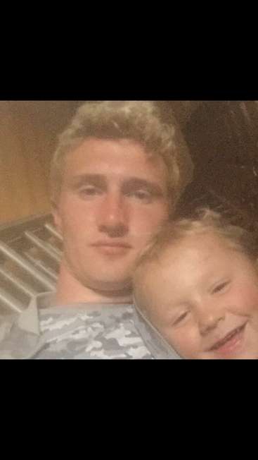 A imagem é uma fotografia em close-up de um homem e um menino, com a cabeça do homem apoiada no ombro do menino. O homem tem cabelo loiro curto.