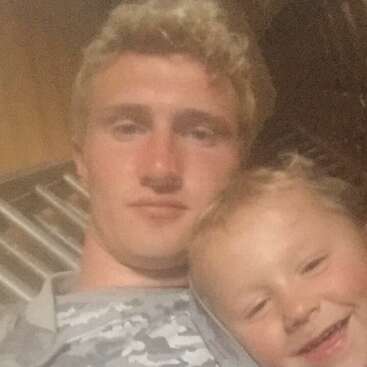 La imagen es una fotografía en primer plano de un hombre y un niño pequeño, con la cabeza del hombre apoyada en el hombro del niño. El hombre tiene el pelo corto y rubio.