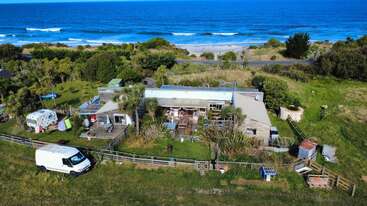 This image shows a coastal house surrounded by greenery, trees, and a fenced yard. There’s a white van, camper, sheds, and the ocean in the background.