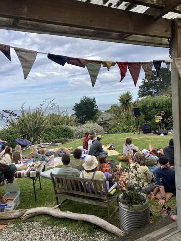 People relax on blankets and benches, watching a musician perform outdoors. Colorful bunting hangs overhead, lush greenery surrounds, and the ocean is visible in the distance.