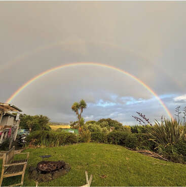 A vibrant double rainbow arcs across a cloudy sky above a lush green garden with palm trees, wooden chairs, and a rustic fire pit, creating a serene scene.
