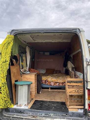 The image shows the back of a van converted into a cozy living space with a colorful bed, wooden furniture, storage crates, and various textiles hanging inside.