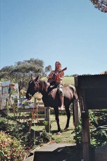 A shirtless man with long hair sits on a large dark horse, playing guitar in a garden, surrounded by lush plants under a bright blue sky.