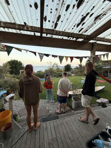 Four people stand on a porch under a weathered roof, looking out at a garden and ocean. Bunting hangs, surfboards and summer vibes abound.
