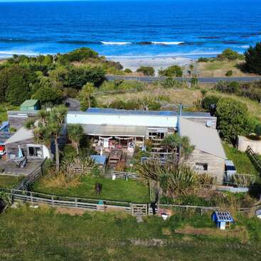 This image shows a coastal house surrounded by greenery, trees, and a fenced yard. There’s a white van, camper, sheds, and the ocean in the background.