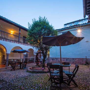 The image depicts a serene courtyard with a tree, tables, chairs, and umbrellas, surrounded by a building with archways and a balcony, evoking a peaceful atmosphere.