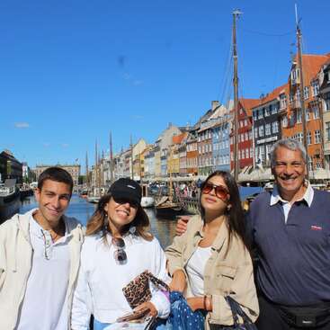 Cuatro personas posan junto a un pintoresco canal en una ciudad europea, con edificios de colores, barcos y un cielo azul despejado, disfrutando juntos de un día soleado.