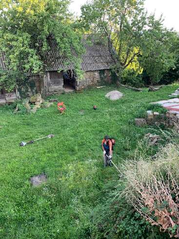Uma pessoa está usando um aparador para cortar grama em um quintal verde e exuberante, com um antigo celeiro de pedra e grandes árvores ao fundo.