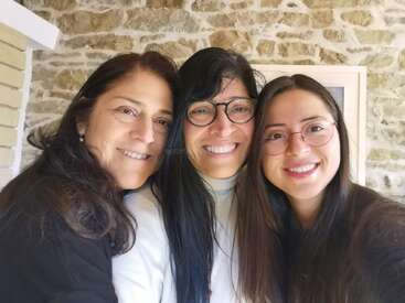 Três mulheres sorrindo juntas, demonstrando cordialidade e felicidade. Elas têm cabelos longos e escuros, usam óculos e parecem estar em uma sala aconchegante com paredes de pedra.