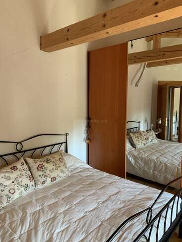 Um quarto aconchegante apresenta uma cama de ferro forjado com um edredom branco e travesseiros florais, vigas de madeira no teto e um guarda-roupa espelhado que reflete o espaço.