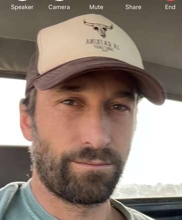 Un homme barbu portant une casquette de camionneur et une chemise décontractée est assis dans un véhicule. L'interface de l'appel vidéo apparaît en haut avec les options haut-parleur, caméra, sourdine, partage et fin.
