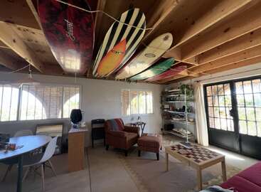 Cette chambre confortable comprend des planches de surf suspendues à un plafond en bois, des meubles confortables, une table basse en damier, de la lumière naturelle, de grandes fenêtres et une étagère bien rangée.