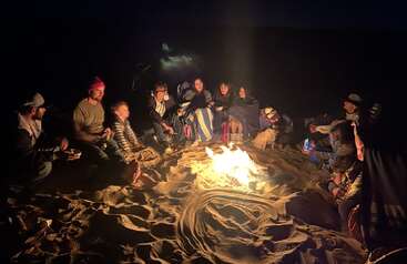 Un groupe de personnes est assis autour d'un feu de camp sur le sable, la nuit, partageant chaleur, histoires et rires ; des couvertures couvrent certains tandis que d'autres profitent de la lueur.